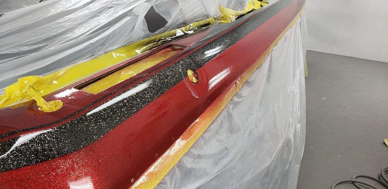 A close-up view of a shiny, metallic red automotive bumper being prepared with masking tape in a workshop.