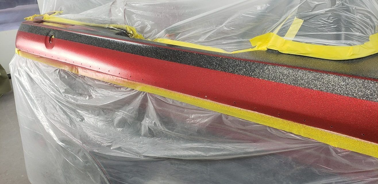 A red boat hull edge with textured black trim, masked off with yellow painter's tape and protected by clear plastic film.