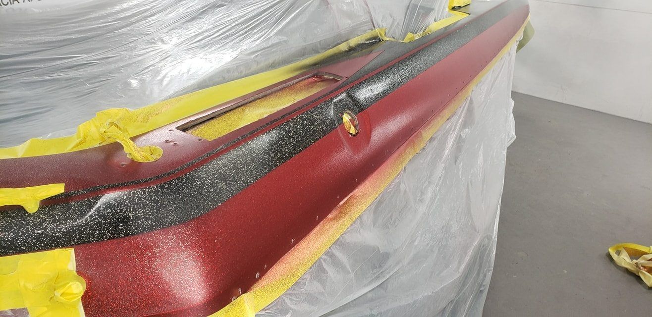 A red automotive part with a textured black stripe, taped off with yellow masking tape in a workshop setting.