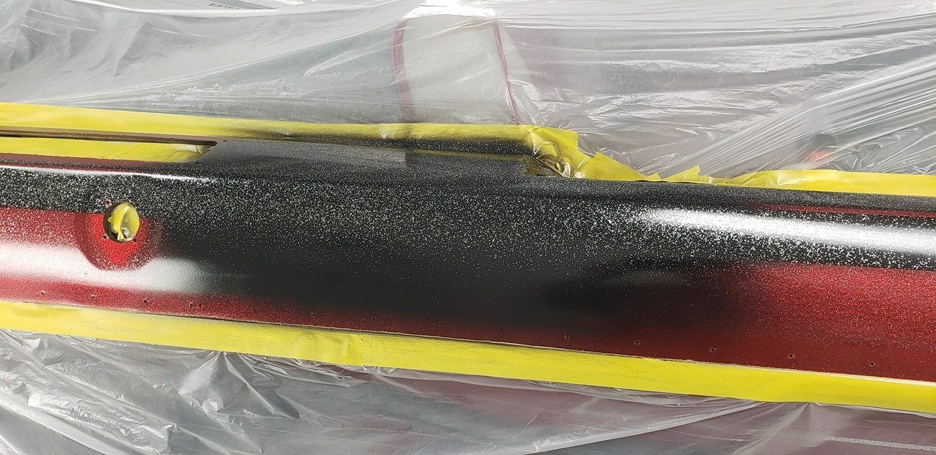 A red car panel masked with yellow tape, partially covered by a textured black spray-on protective coating.