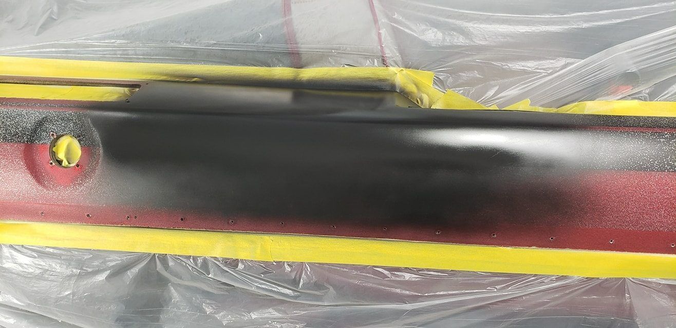 A car part being repaired, with a black-painted area masked by yellow tape against a red metallic surface.
