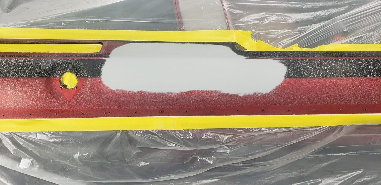 A closeup of a car bumper taped off with yellow masking tape, featuring a sanded gray primer patch over red and black paint.