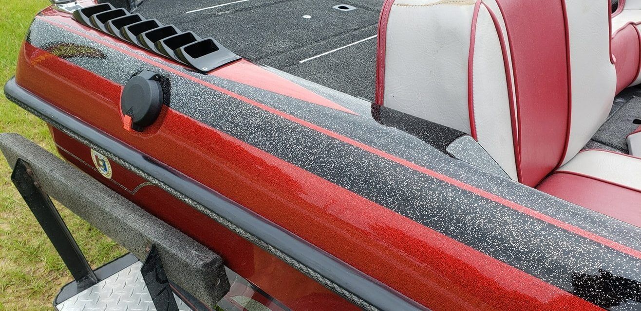 A close-up of a red and black speckled fiberglass bass boat gunwale with black rod holder slots and gray and red seats.