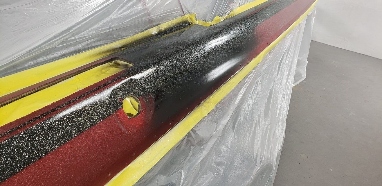 A close-up view of a long, metallic bar being spray-painted with black paint, masked off with yellow tape on a plastic cover.