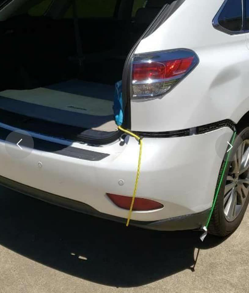 A white SUV with a detached rear bumper held in place by yellow and green ropes attached to the vehicle frame.
