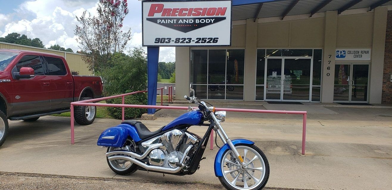 A bright blue custom motorcycle parked in front of Precision Paint and Body shop with a red truck visible on the left.