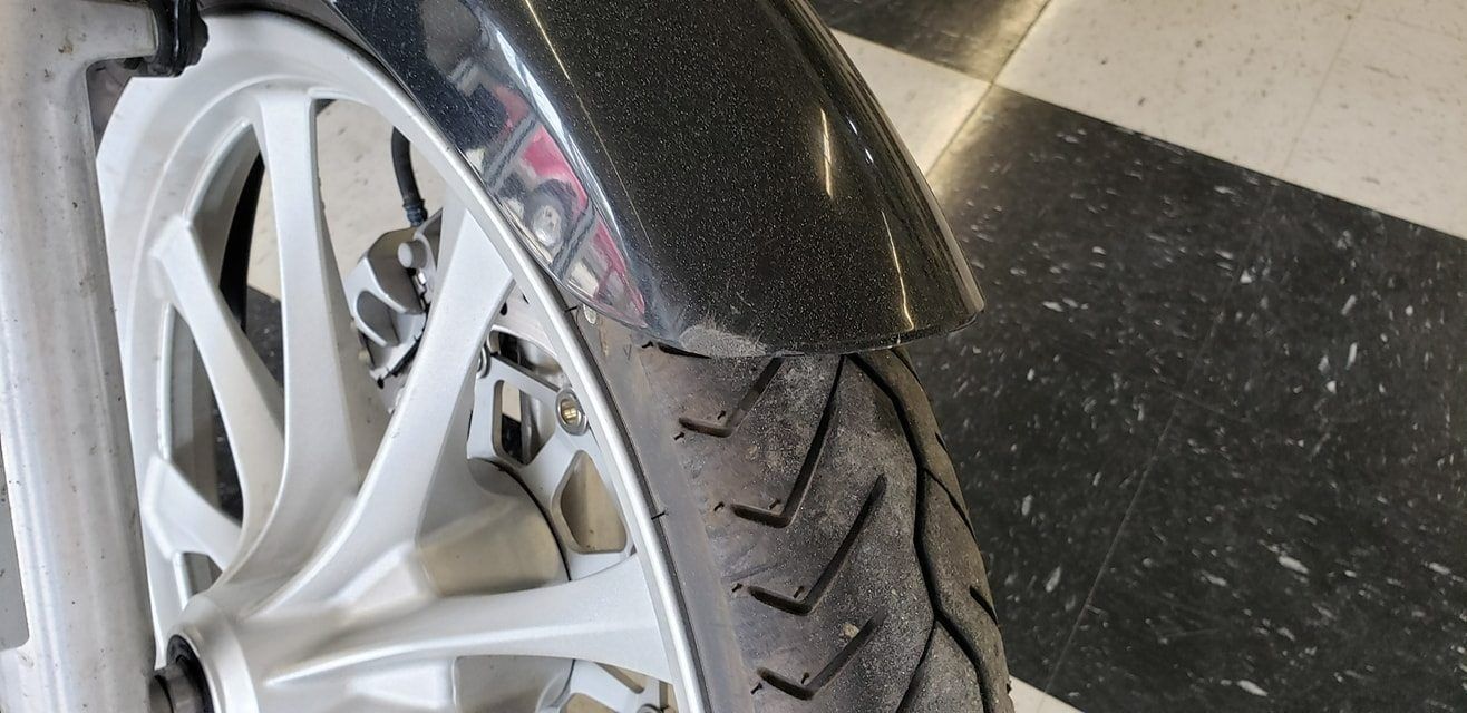 A close-up of a motorcycle's front wheel with white spokes, a textured black fender, and a patterned tire.