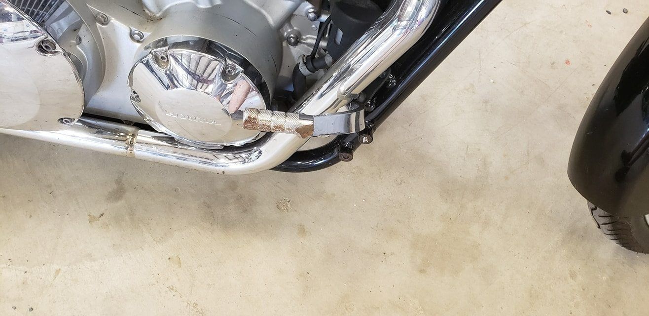 A close-up of a motorcycle engine showing a chrome side cover with a stained, metal kickstand arm extending outward.