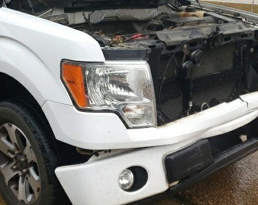 Close-up of a damaged white Ford pickup truck with its hood open, showing impact damage to the front bumper and grille.