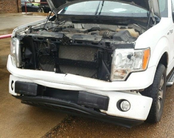 A white pickup truck with a damaged front bumper and missing grille parked outdoors with the hood open.
