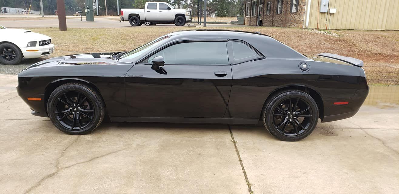 A black Dodge Challenger parked on a paved lot with a silver pickup truck in the background.