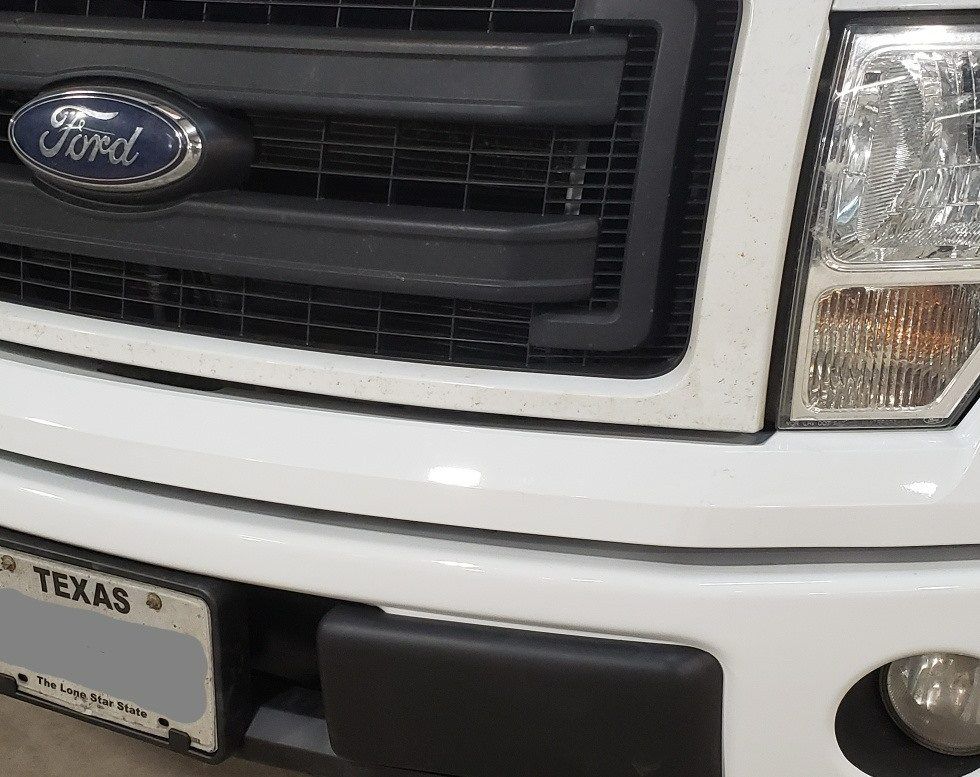 Close-up of a white Ford truck's front grill, emblem, headlight, bumper, and partial Texas license plate.