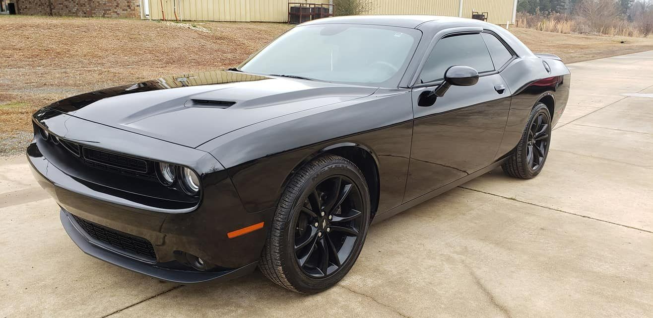 A black Dodge Challenger parked on a concrete driveway against a rural backdrop.