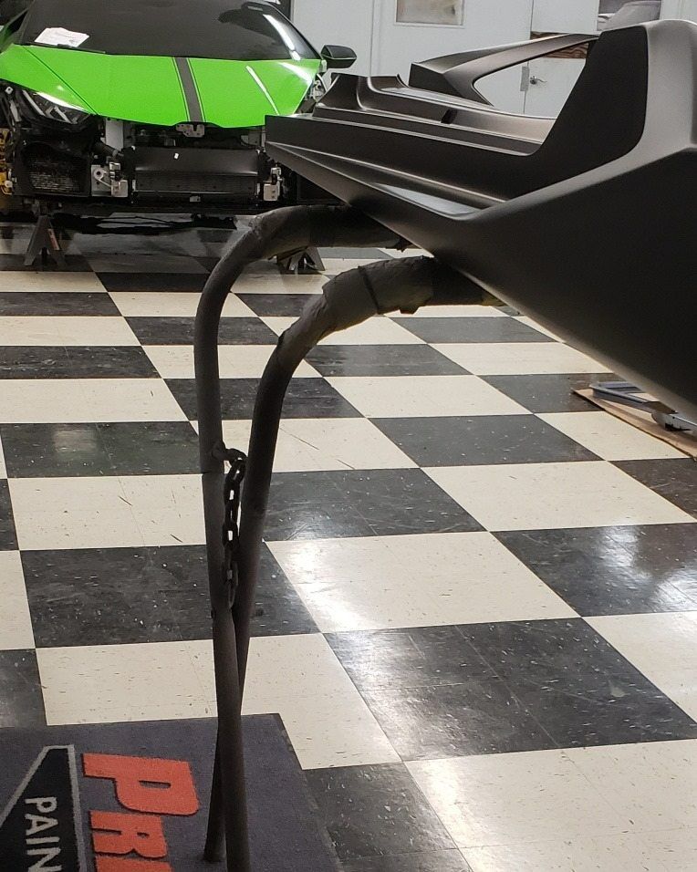 A black car bumper part sits on a metal stand in a workshop with a checkered floor, in front of a green Lamborghini.