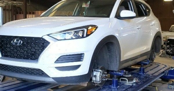A white Hyundai Tucson SUV is raised on a metal car lift in an auto repair shop with its front wheels removed.