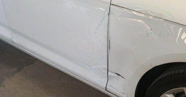 A close-up of a white car with significant denting and paint damage on the front fender and passenger-side door.