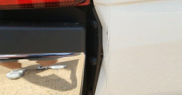 Close-up of a damaged, white car bumper panel with a visible vertical crack, separated from the black rear trim.