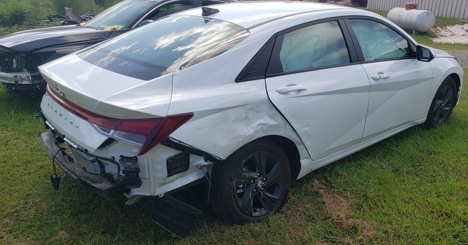 A damaged white sedan sits on grass, missing its rear bumper with visible dents on the rear passenger side quarter panel.