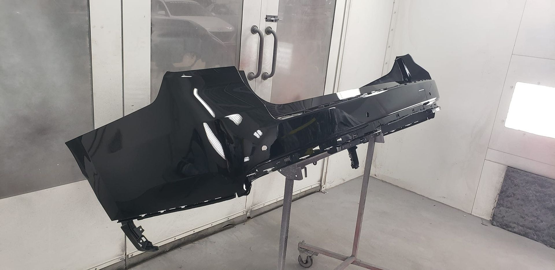 A black, primed automotive bumper panel sits on a metal stand inside a white painting booth.