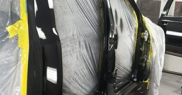 A vehicle’s door frame and interior pillar being prepped for paint, masked with plastic sheeting and yellow tape.
