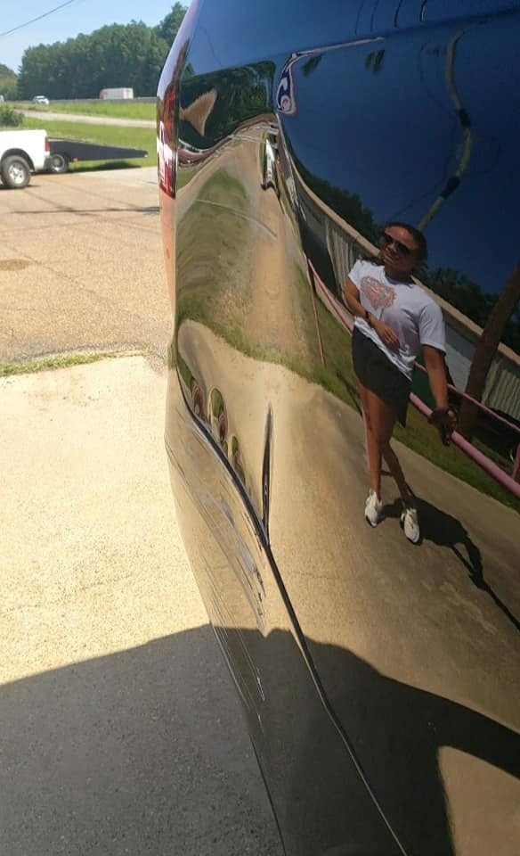 A reflective surface of a vehicle captures a person standing and posing on a sunny day in an outdoor parking area.