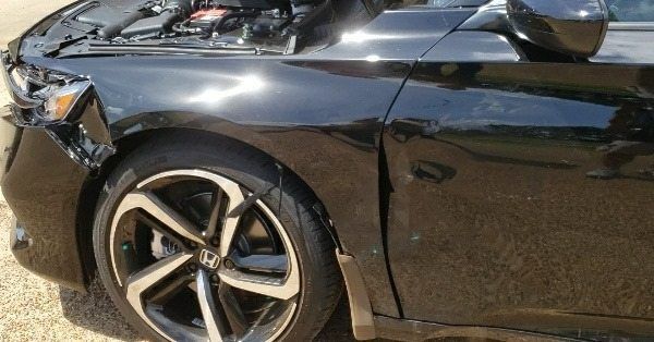 A damaged black Honda car showing a crushed front bumper, detached headlight, and a dented front driver-side panel.