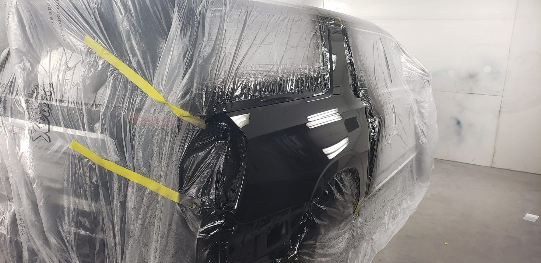 A black vehicle partially covered in protective plastic film inside an auto body repair shop.