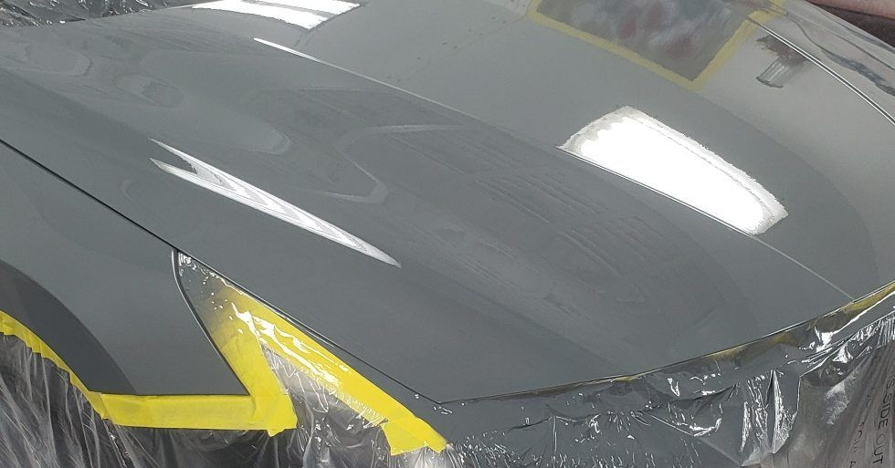 A gray car hood being prepped for paint, with yellow masking tape applied along the edges.
