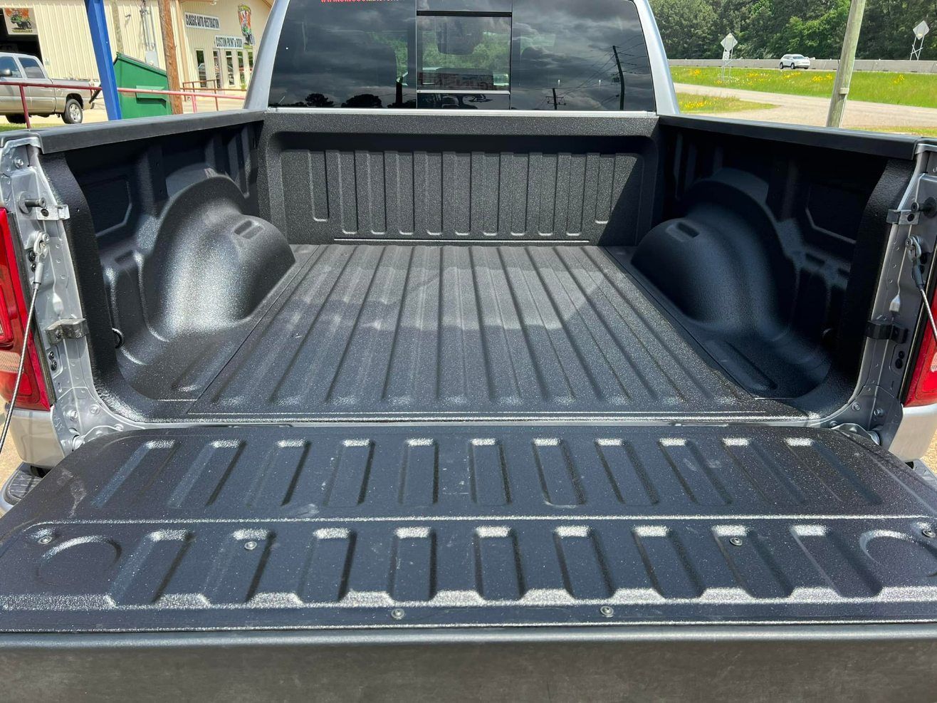The open cargo bed of a silver pickup truck with a black spray-in bed liner, viewed from behind with the tailgate down.