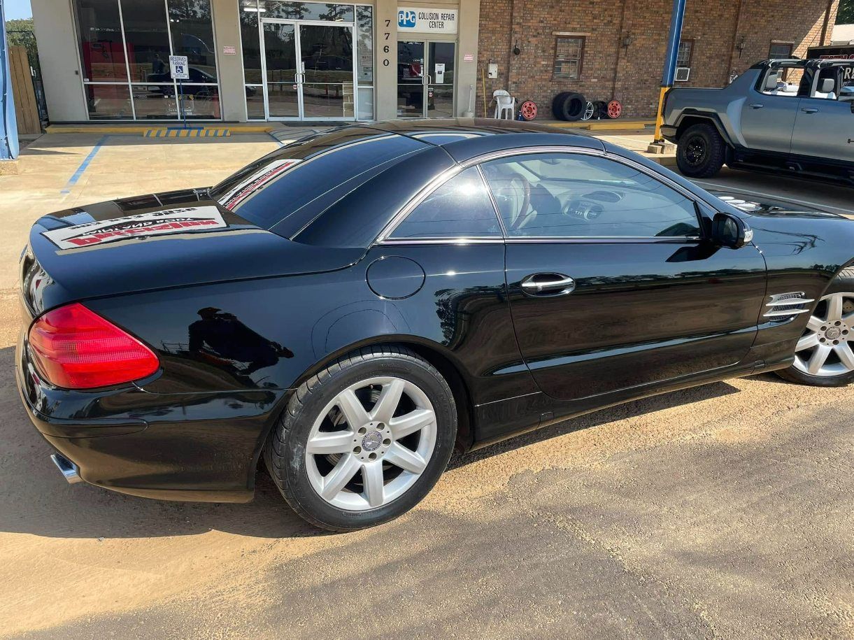 A black Mercedes-Benz SL convertible parked on a gravel lot in front of a brick building.