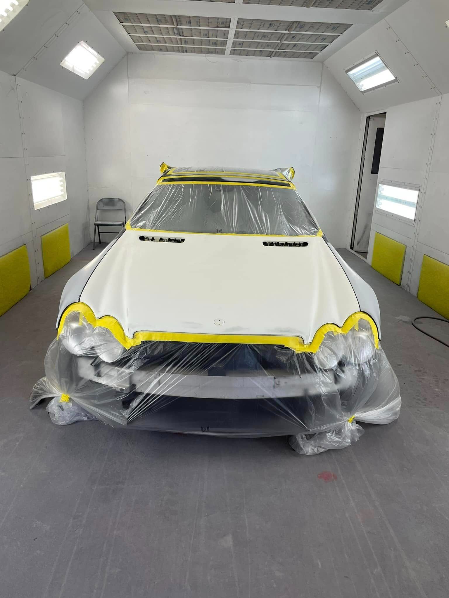 A white car in a spray booth, with the windshield and front masked with plastic and tape for a paint job.