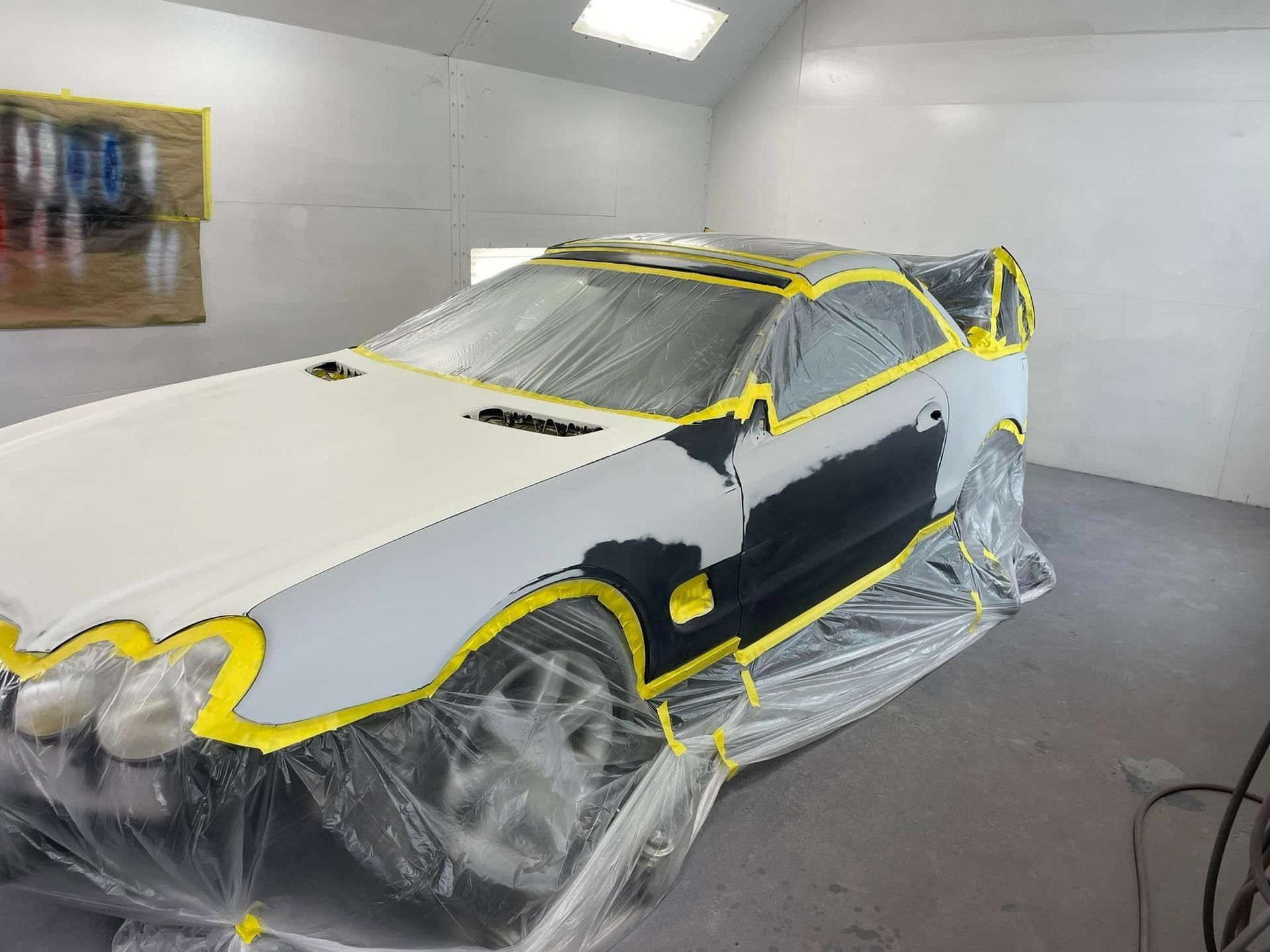 A car in a paint booth covered in plastic masking and tape, with parts of the body prepped for painting.