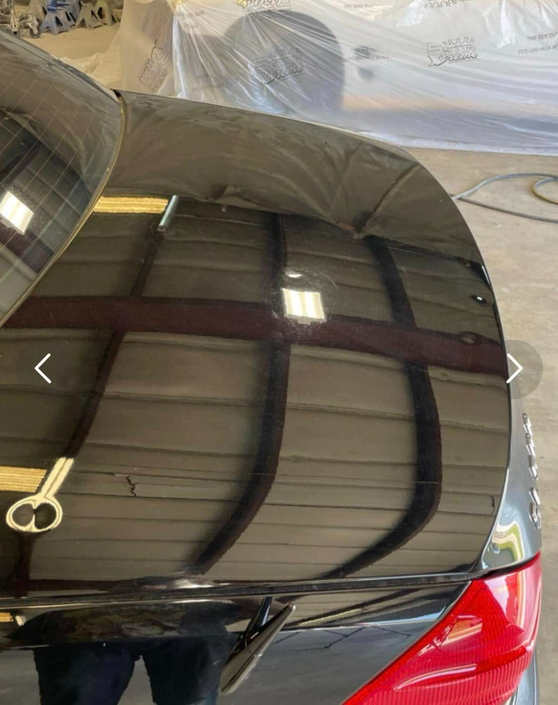 The trunk of a glossy black car reflecting interior ceiling lights and structural beams in a workshop setting.