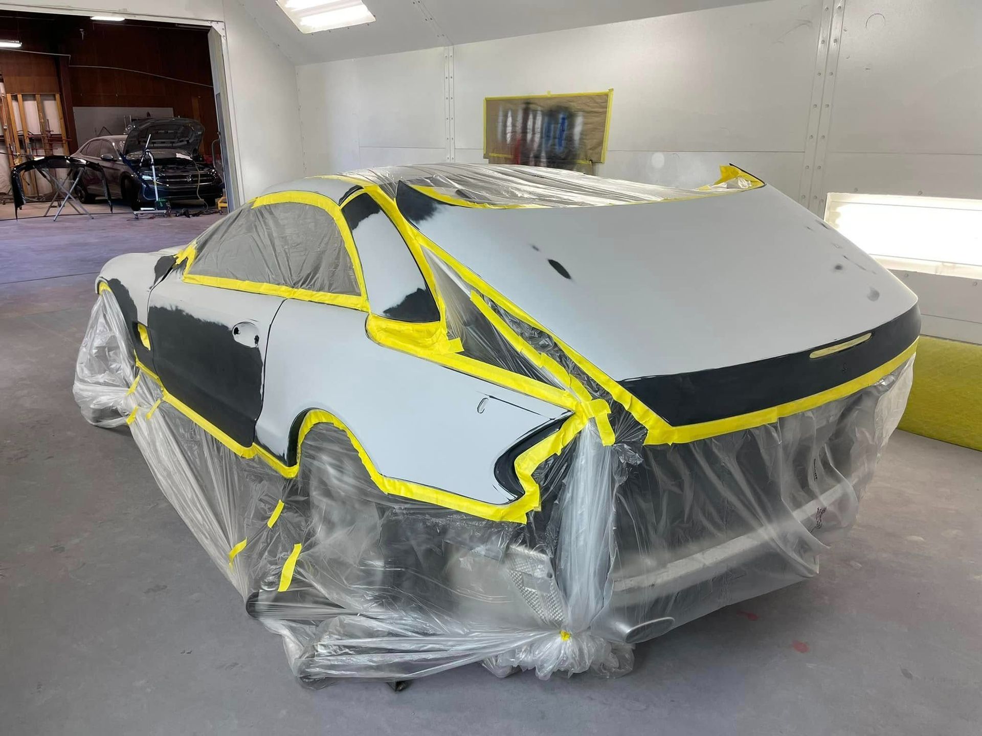 A car covered in protective plastic and yellow painter's tape, undergoing prep work inside an auto body shop.