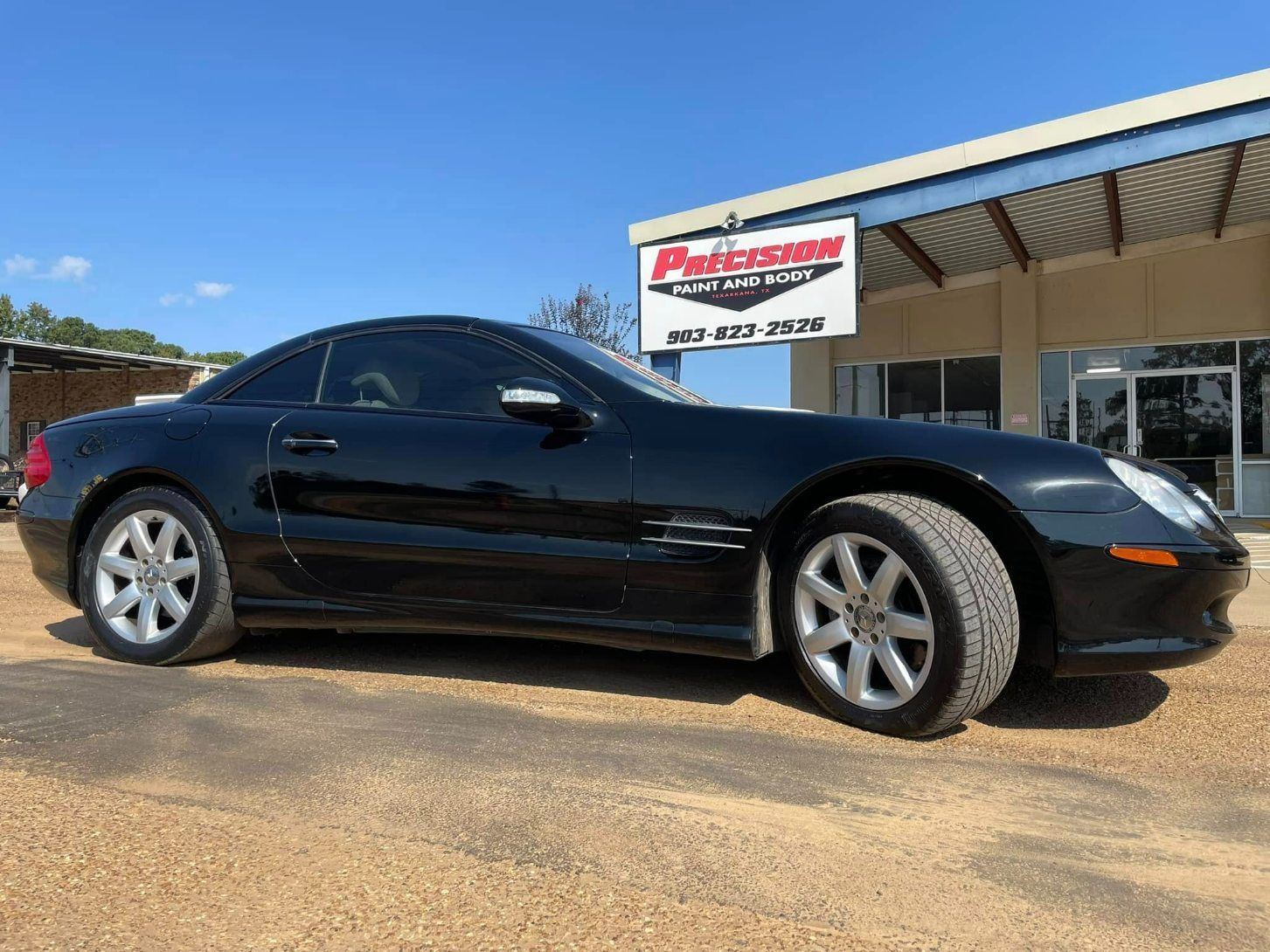 A black Mercedes-Benz SL-Class convertible parked on a gravel lot in front of a dealership building under a blue sky.