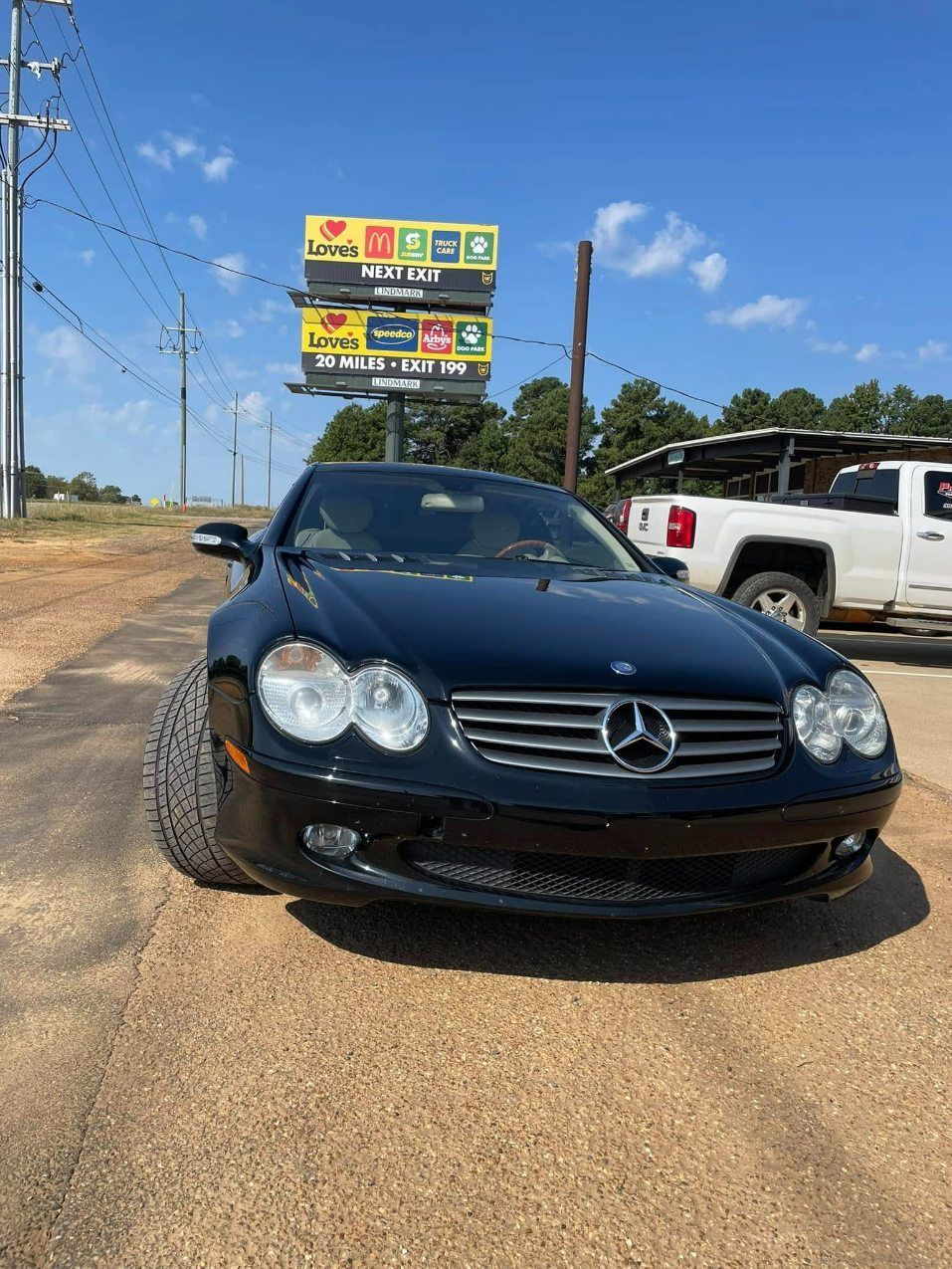 A black Mercedes-Benz parked on a gravel lot under a bright blue sky, with a commercial sign and white truck in the back.