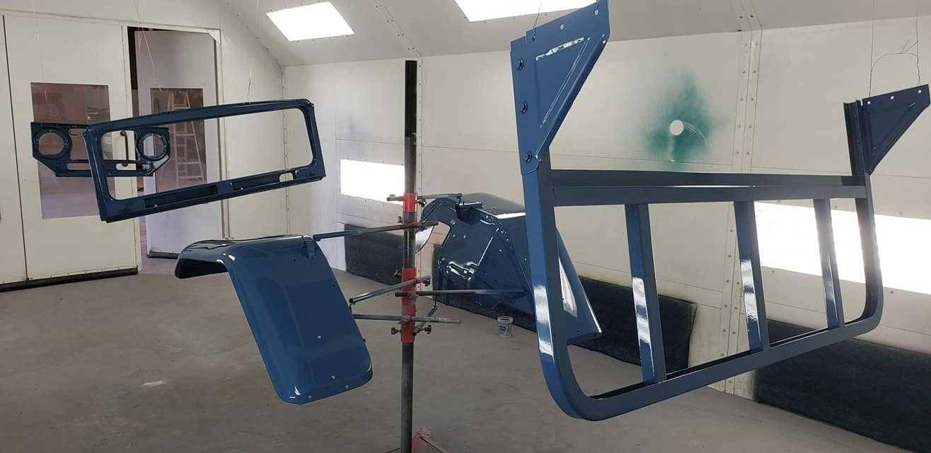 Blue-painted metal vehicle body parts hang to dry in a bright, white-walled spray booth.