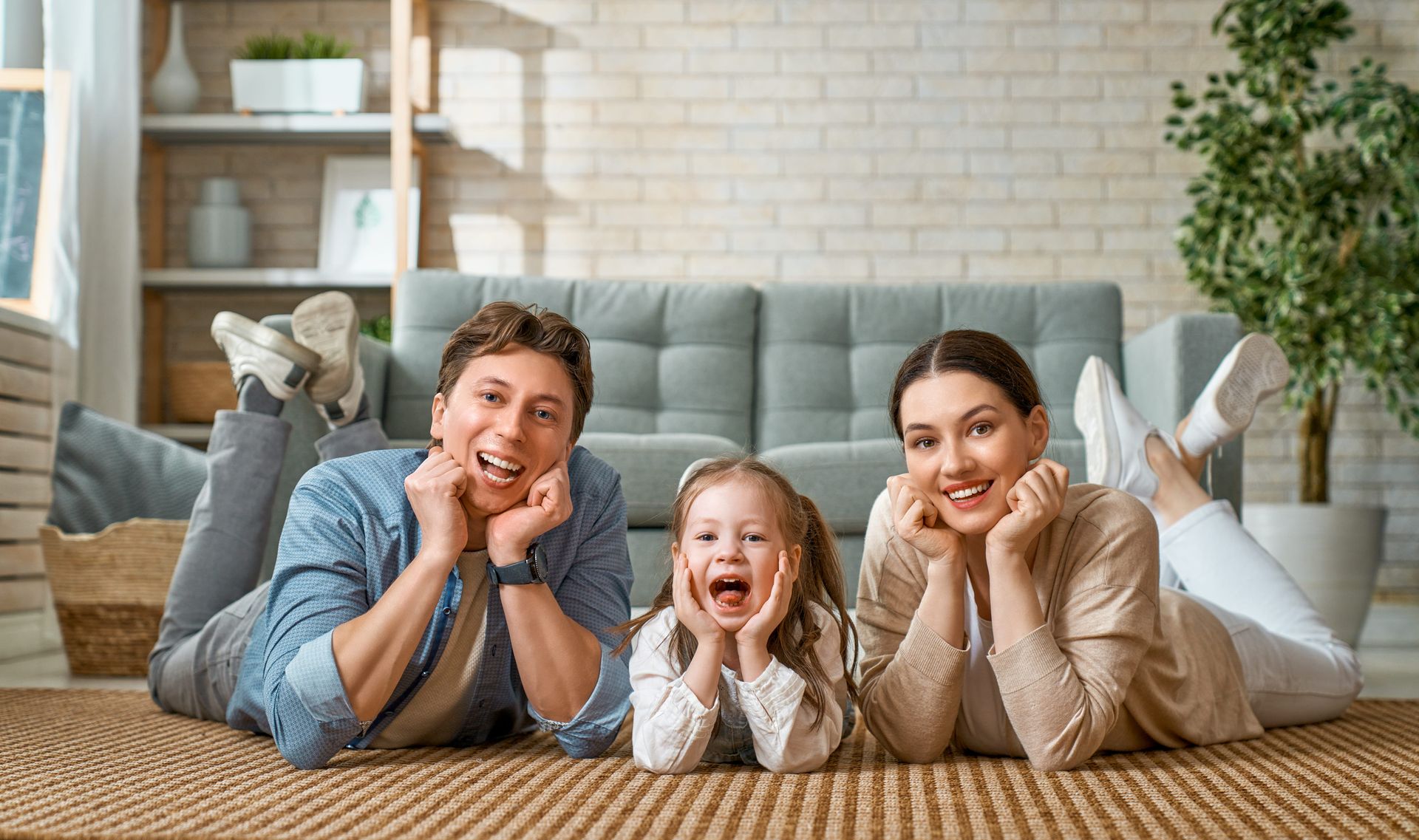 Three people lying on the floor, smiling in a cozy living room with a couch and plants.