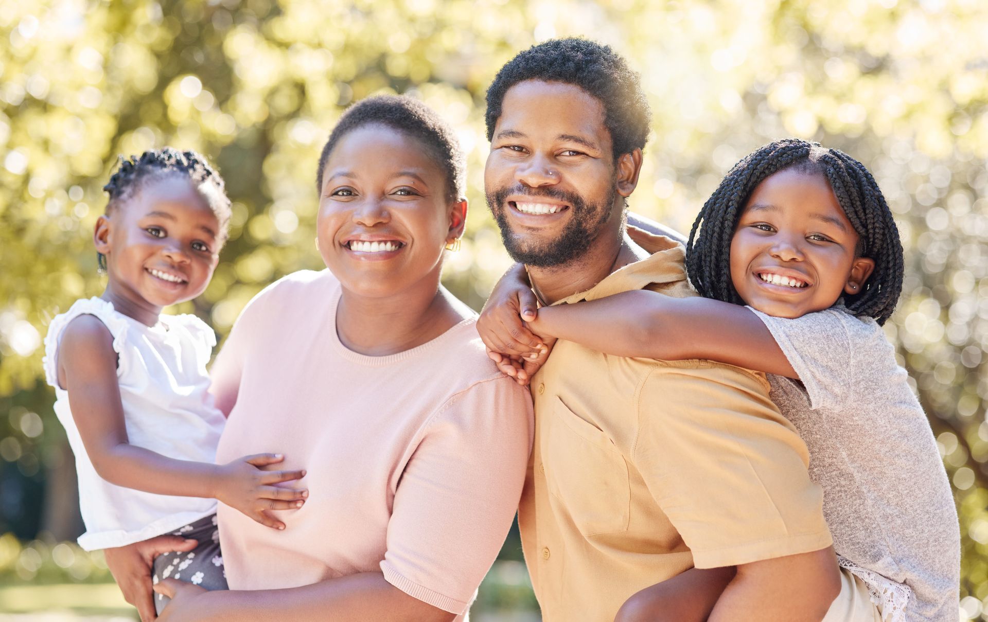 Smiling family of four outdoors, with two children hugging their parents in warm sunlight.