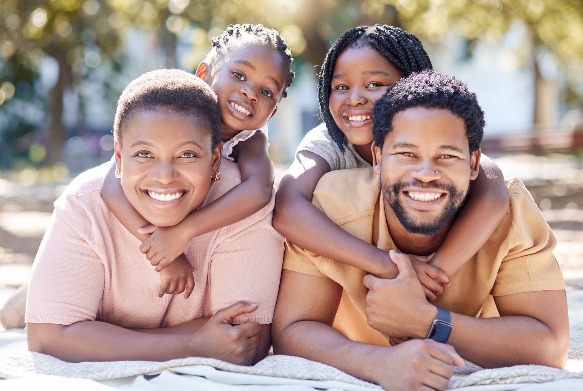 Two smiling couples lying on the beach, with one child on each adult’s back in bright sunlight