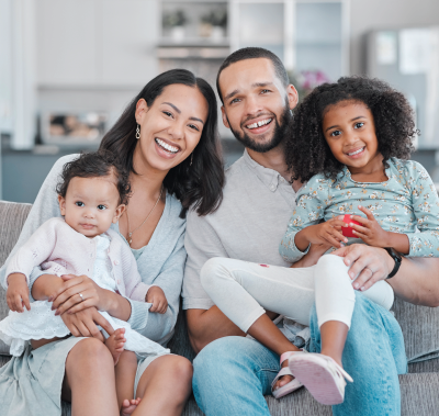 Smiling family of four sitting on a couch in a bright living room, holding two young children.