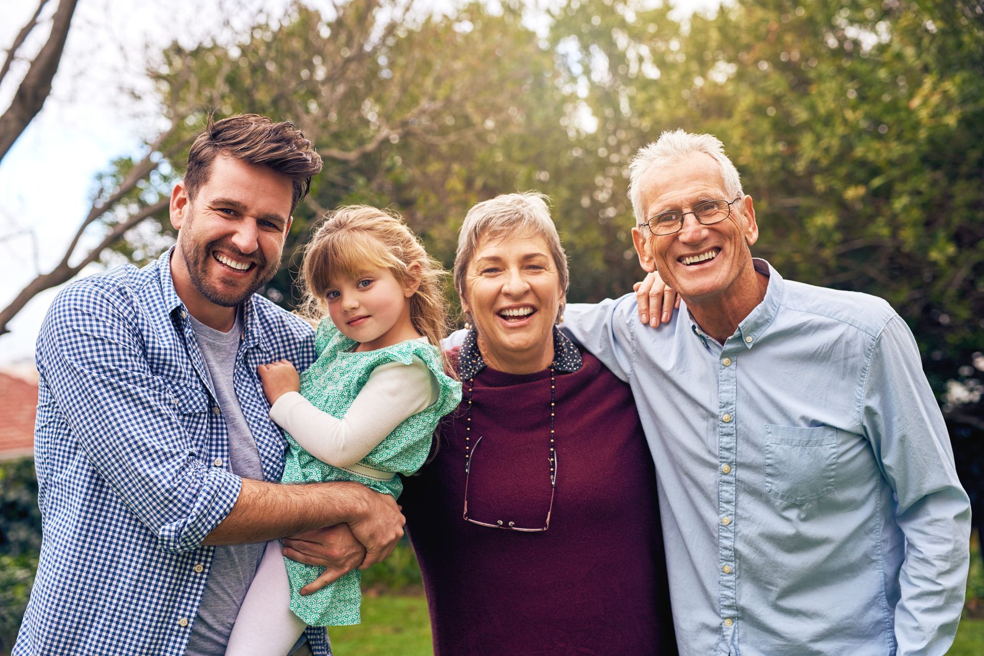Four smiling people standing outdoors in a park, with trees and greenery in the background.