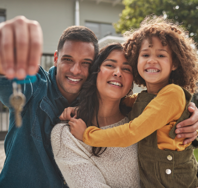 Three people smiling outdoors, hugging near a house while someone holds up keys in the foreground