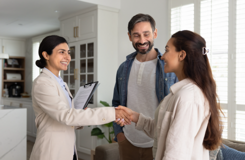 Two people shake hands as a woman with a clipboard smiles in a bright living room