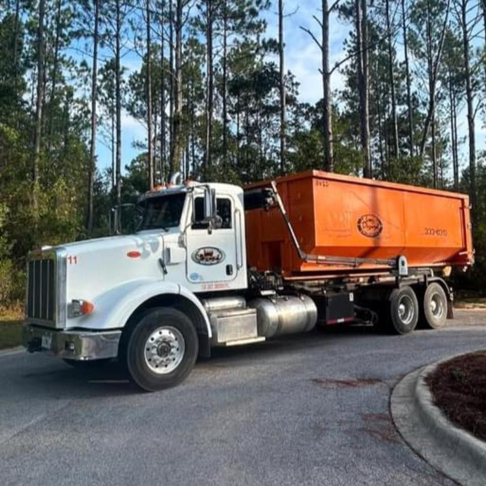 White semi-truck with orange dump trailer on a paved road beside tall pine trees. White semi-truck with orange dump trailer on a paved road beside tall pine trees.