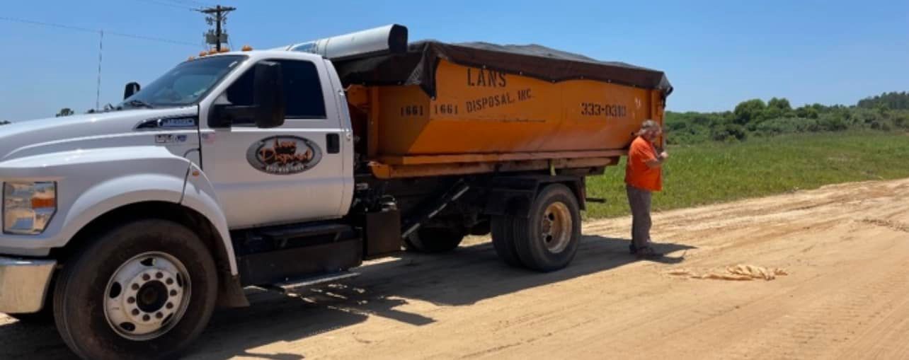 White dump truck with orange bed parked on a dirt road beside a worker in orange safety gear