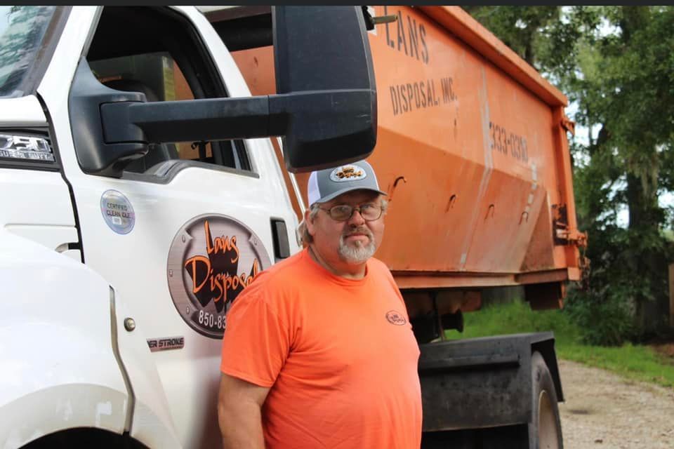 Man in orange shirt and cap standing beside a white truck with an orange dump bed outdoors
