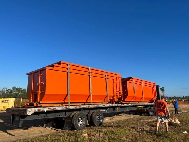 Orange utility containers on a flatbed trailer in a dirt lot, with two people nearby under a blue sky Orange utility containers on a flatbed trailer in a dirt lot, with two people nearby under a blue sky