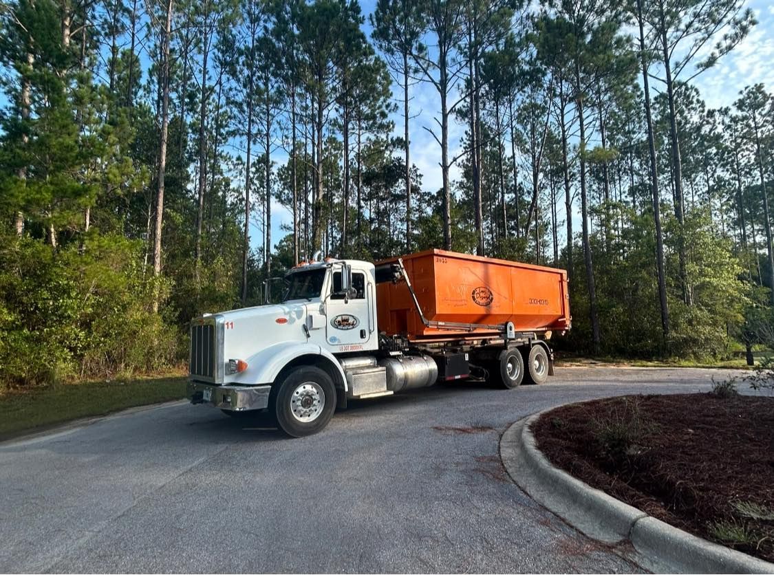 White dump truck with orange trailer driving on a curved road beside tall pine trees.