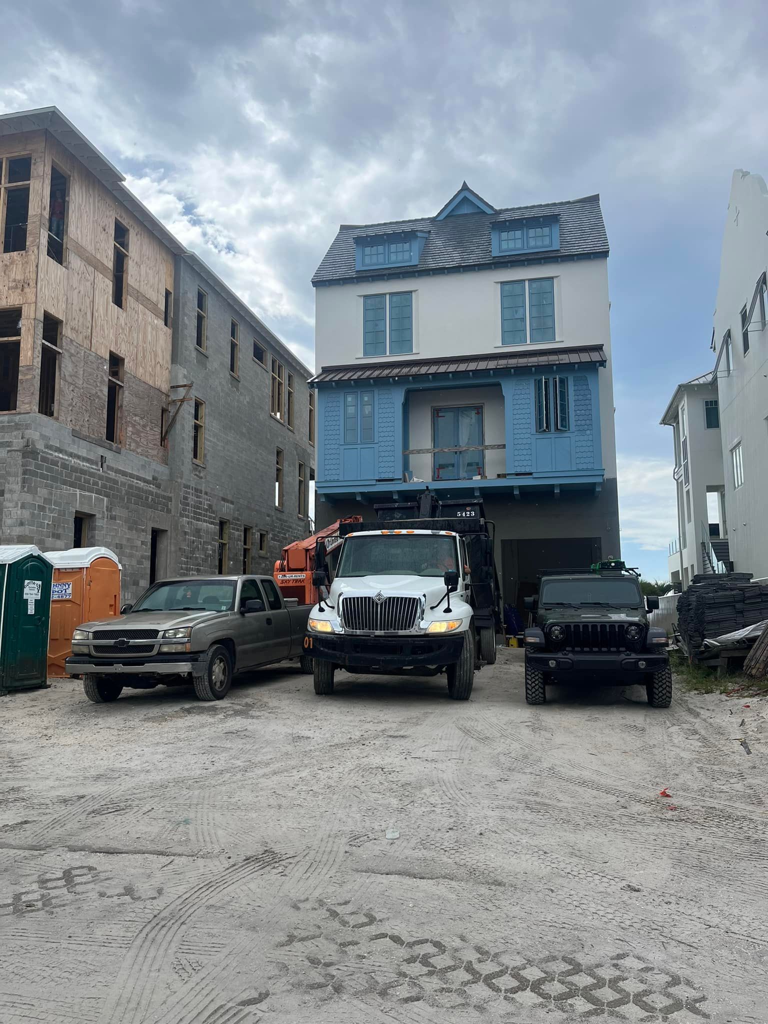 Blue house under construction with parked vehicles on a gravel lot, flanked by unfinished buildings